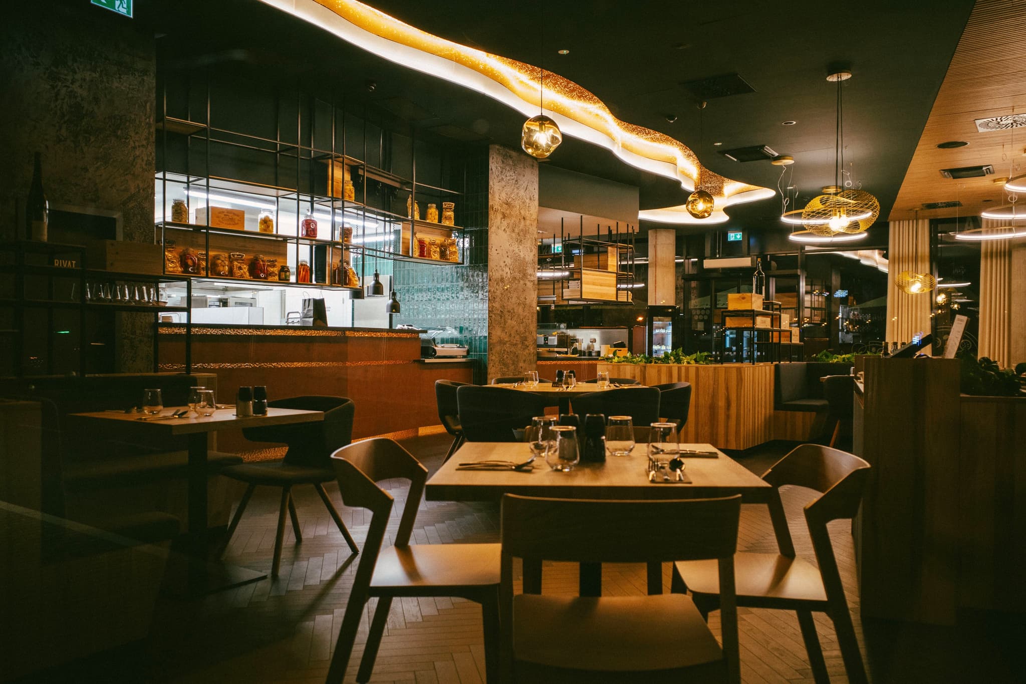 A dimly lit, modern restaurant interior with wooden tables and chairs, ambient lighting, and a bar area in the background