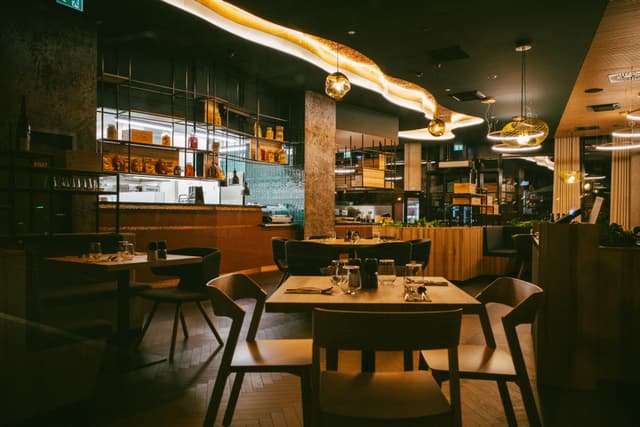 A dimly lit, modern restaurant interior with wooden tables and chairs, ambient lighting, and a bar area in the background