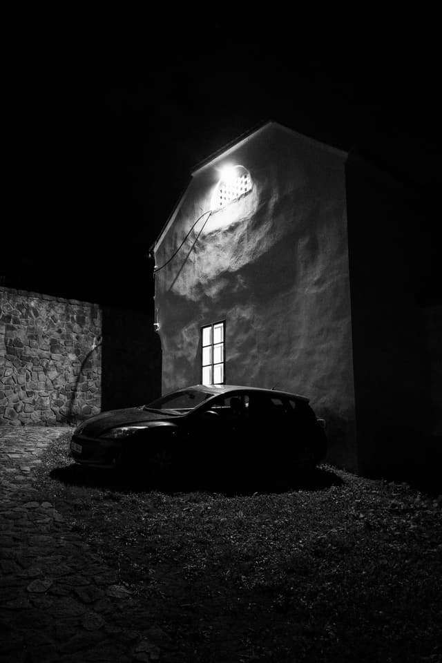 A black and white scene of a car parked beside a building at night, with a light illuminating the building's wall and window
