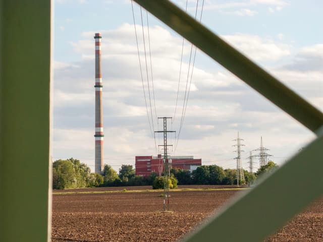 A tall industrial chimney and power lines in a rural landscape, with a red building and trees in the background, framed by a green metal structure