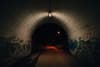 A dimly lit tunnel with graffiti on the walls, leading to a dark path illuminated by a distant streetlight