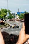 A Formula 1 car is racing on a city street, surrounded by a large crowd of spectators Trees and buildings are visible in the background, and people are capturing the moment with their phones