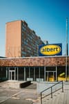 A building with a large albert sign in front, featuring a brick facade and a multi-story apartment block in the background under a clear blue sky