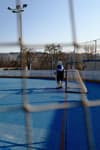 A hockey goalie stands in front of a net on an outdoor rink, viewed through a protective netting. The rink has a blue surface, and there are trees and buildings in the background under a clear sky