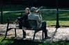 Two people sitting on a park bench, one smoking, with a grassy background and dappled sunlight