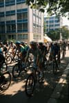 A group of cyclists riding through an urban area with buildings in the background, shaded by trees