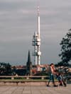 A tall, modern tower stands prominently against a cloudy sky, with a church spire and rooftops in the foreground A person walks with a child in a stroller on a paved area, and a tree is visible on the right