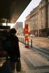 A person with a backpack stands at a bus stop on a sunlit street, with buildings and a red bus stop sign in the background