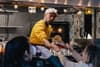 A man in a yellow sweater and cap serves food from a food truck to two women