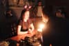 A dimly lit room with a young woman wearing star-shaped headbands, sitting at a table with a lit birthday cake in front of her Another person is leaning over the table, and a few people are in the background